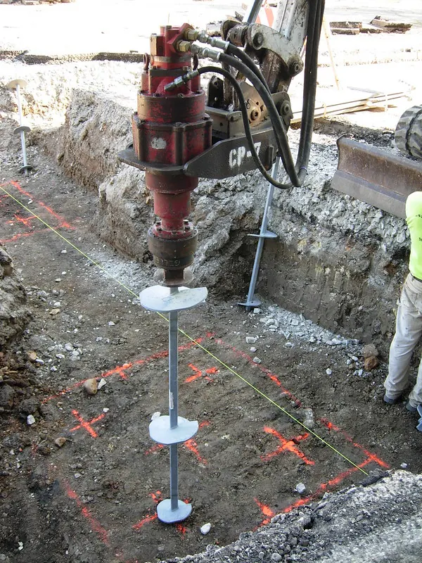 Batang helical pile dengan tiga pelat heliks terhubung ke kepala penggerak hidraulik di area galian